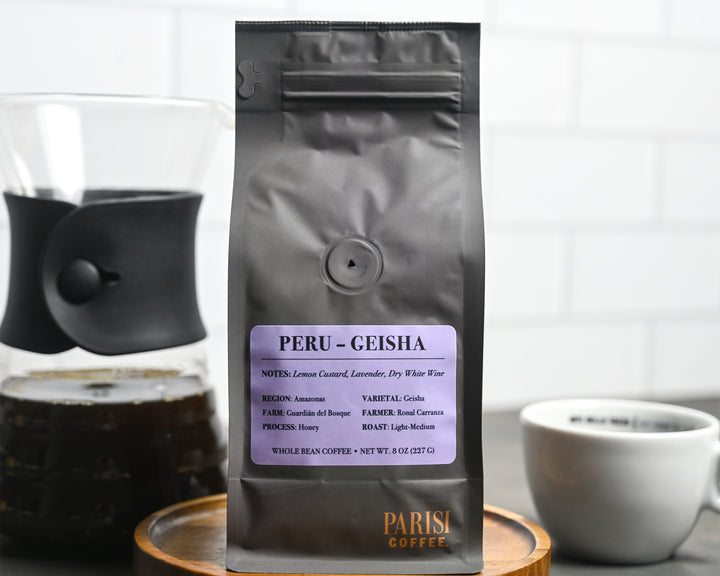 Coffee bag labeled 'Peru Geisha' with a coffee cup and grinder in the background.